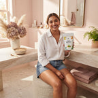 A woman holding a container of Natural Bikini Wax in a bathroom with blush pink and rose-colored decor.